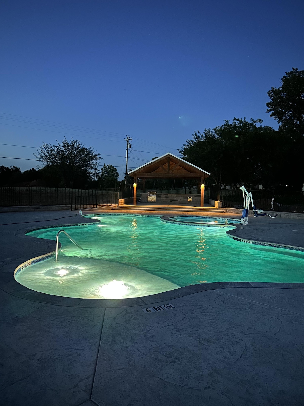 The illuminated pool area is showcased at dusk, featuring a unique shape with glowing lights beneath the water. A covered gazebo can be seen in the background, adding a shaded seating option. Fencing surrounds the pool, providing a secure environment.