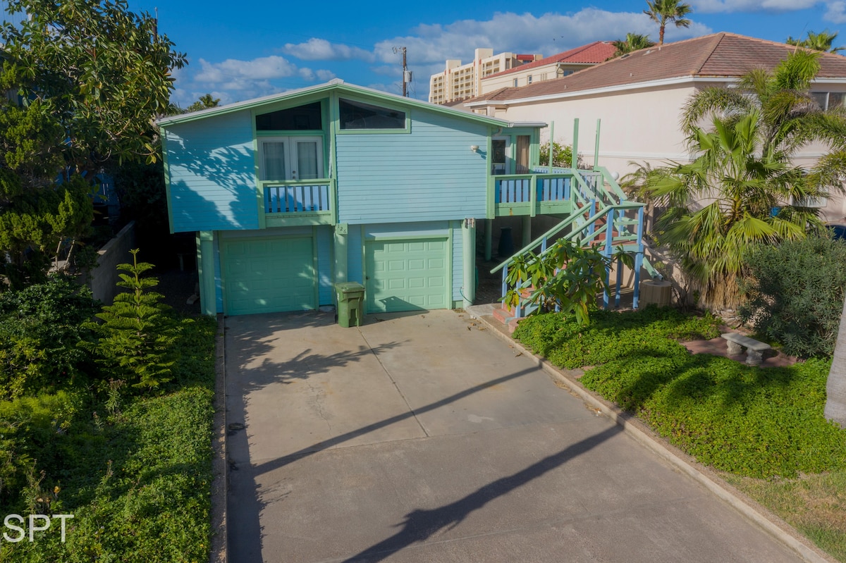 The exterior of a light blue duplex is visible, set amidst lush greenery. A driveway leads to two garage doors, with a large palm tree casting a shadow. A flight of stairs leads to a second-level entrance, providing access to the upper unit.