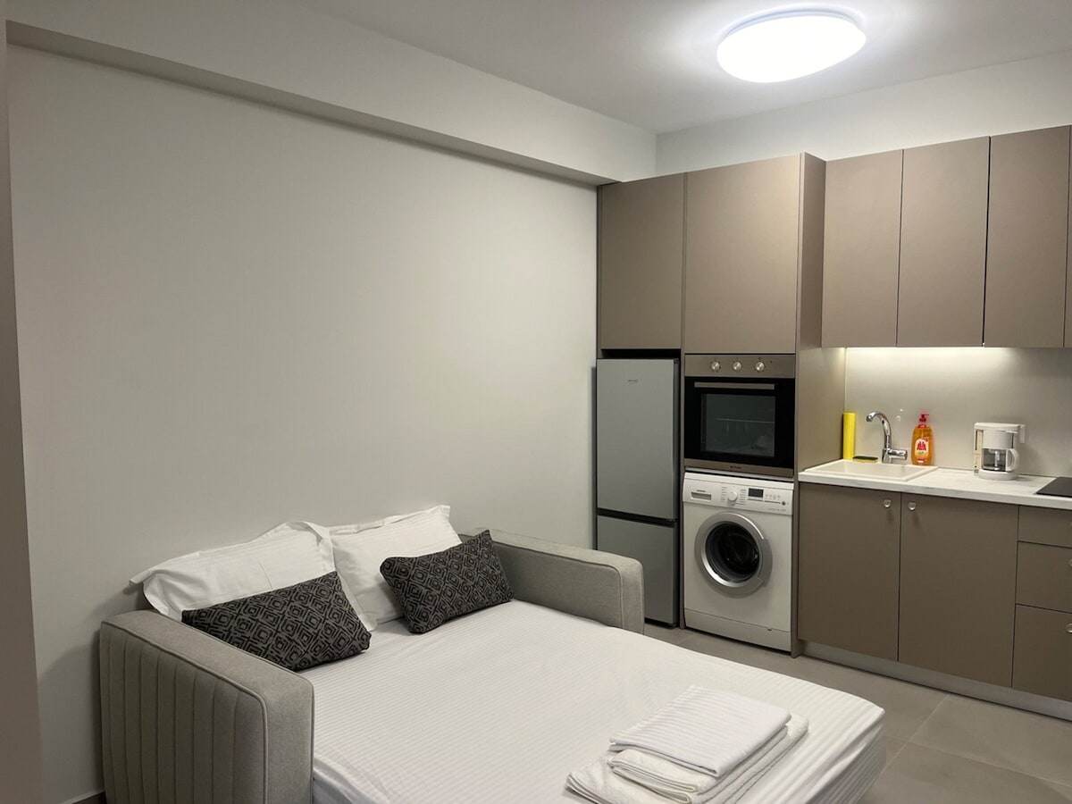 A compact living space showcases a sofa bed with decorative pillows positioned in front of a modern kitchen. The kitchen is equipped with a refrigerator, oven, and washing machine, offering essential appliances for convenience. Soft lighting enhances the functional atmosphere.