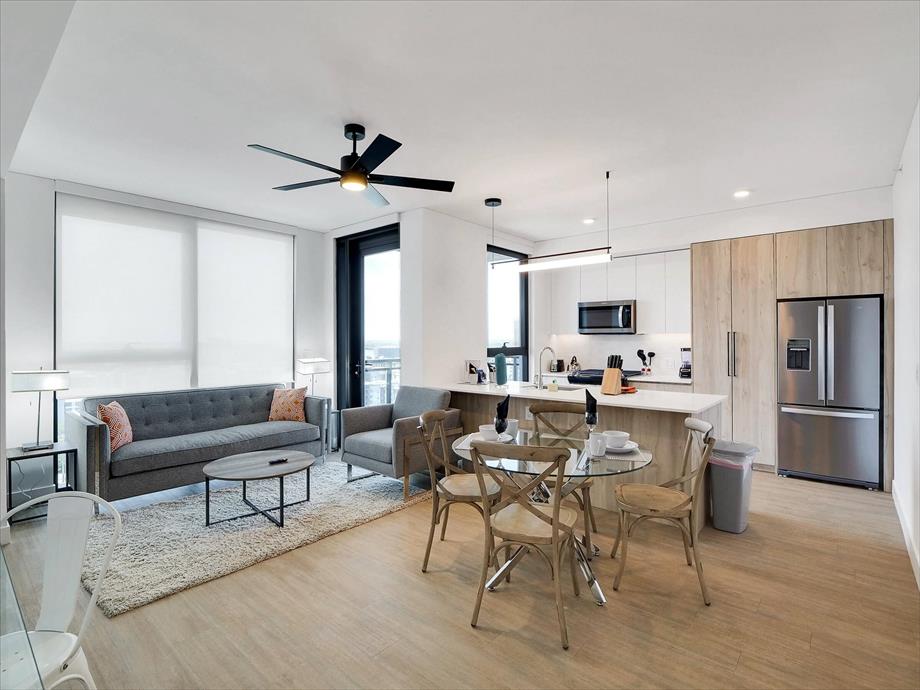 A modern open-concept living space features a gray sofa, circular dining table with four chairs, and a well-equipped kitchen with stainless steel appliances. Large windows allow ample natural light, and a light-colored area rug complements the wooden flooring.