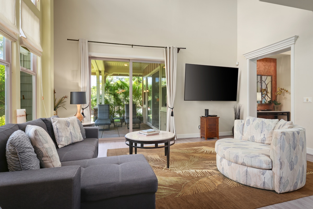A spacious living room is featured, showcasing a modern design with comfortable seating arrangements. Natural light floods the space through large glass doors, leading to a lush outdoor area. A mounted TV and a round coffee table accentuate the inviting atmosphere.