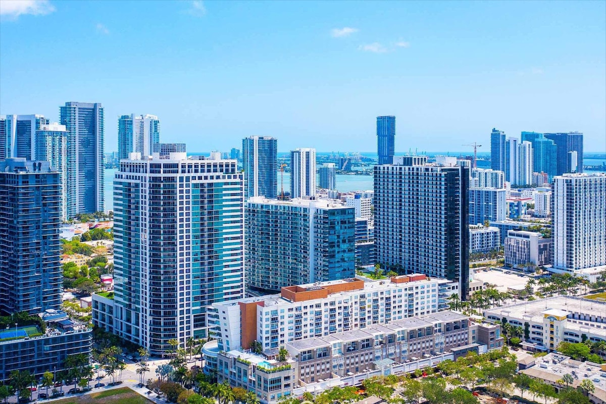 A panoramic view of the Miami skyline is presented, highlighting a mix of modern high-rise buildings and urban architecture. The skyline is set against a bright blue sky, with glimpses of water visible in the background, reflecting the vibrant city atmosphere.