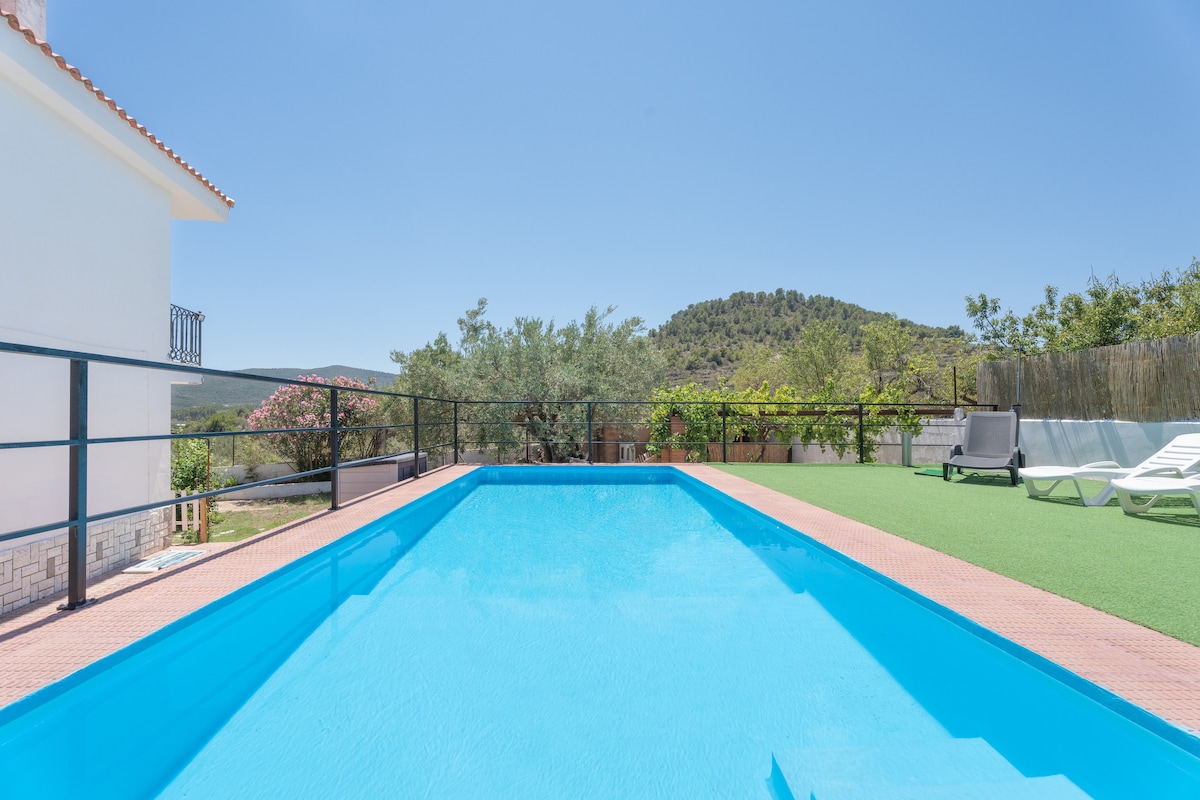 A private outdoor pool is featured, surrounded by a fenced area. Sun loungers are positioned nearby on a grass surface. Lush greenery and a hillside are visible in the background, contributing to a serene environment.