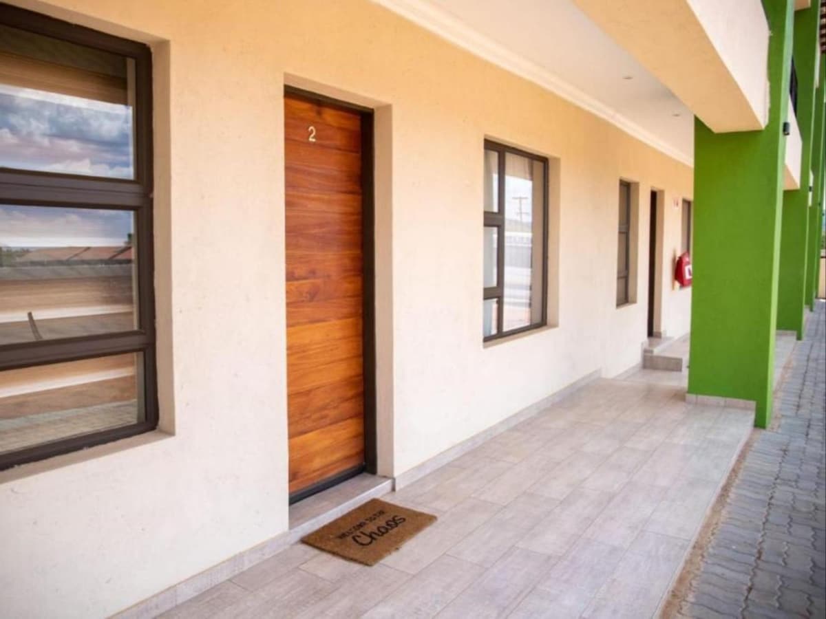 An exterior view of a building featuring light-colored walls and green accents. A wooden door numbered '2' is visible, accompanied by a welcome mat. Large windows provide natural light, and a clean pathway leads to the entrance.