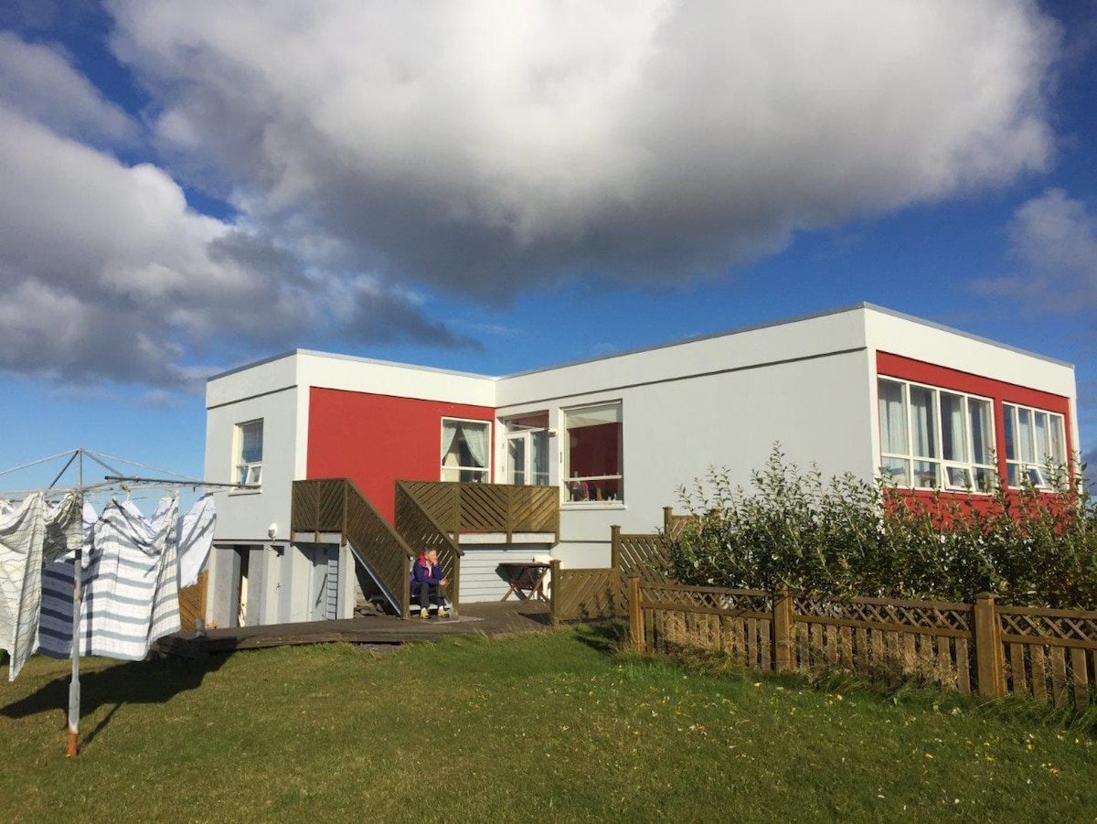 A modern house exterior is presented, featuring a combination of white and red walls. Large windows allow natural light into the living spaces. A grass area surrounds the building, with a clothesline on one side and a wooden fence that encloses the yard.