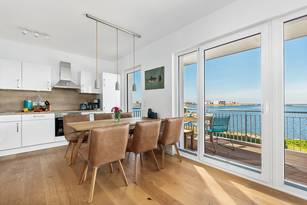 The open-plan kitchen and dining area features a wooden dining table surrounded by leather chairs. Natural light floods in through large glass doors, showcasing a serene water view. A well-equipped kitchen with modern appliances is visible in the background, enhancing functionality.