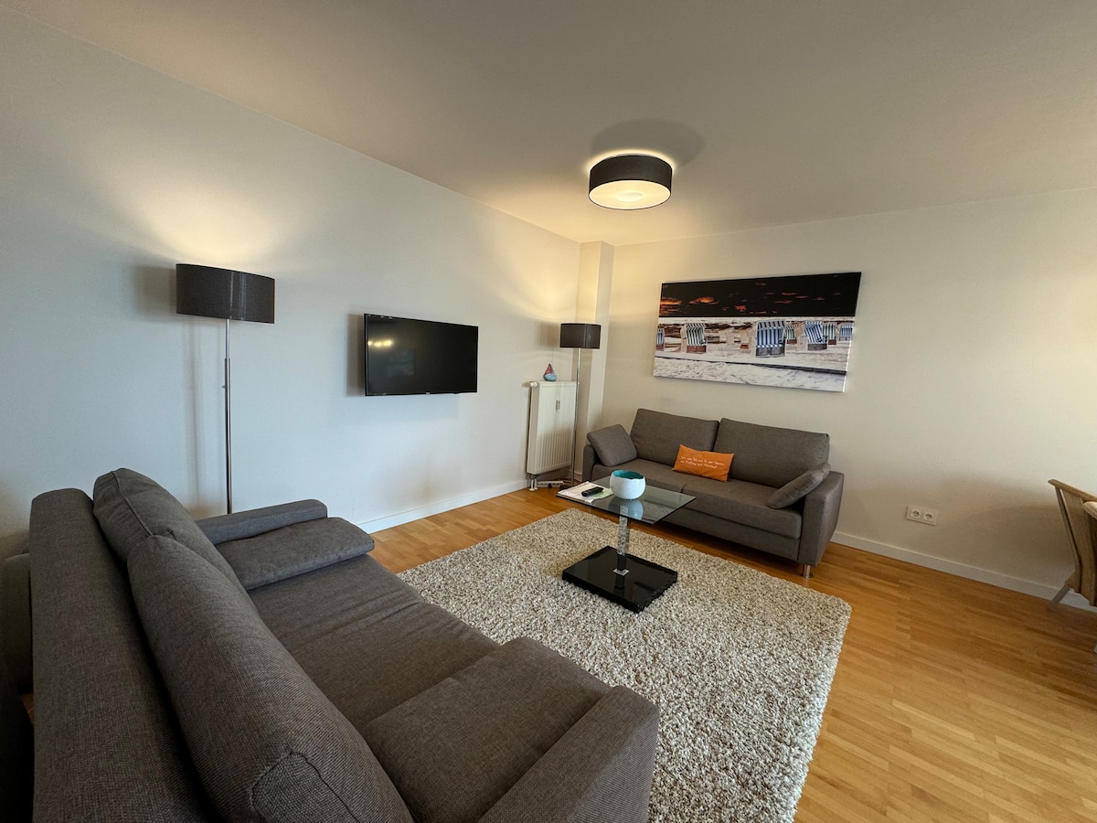A spacious living area features a comfortable seating arrangement with two grey sofas. A flat-screen TV is mounted on the wall, opposite a stylish coffee table, placed on a soft rug. Soft lighting is provided by modern floor lamps, creating a cozy atmosphere.