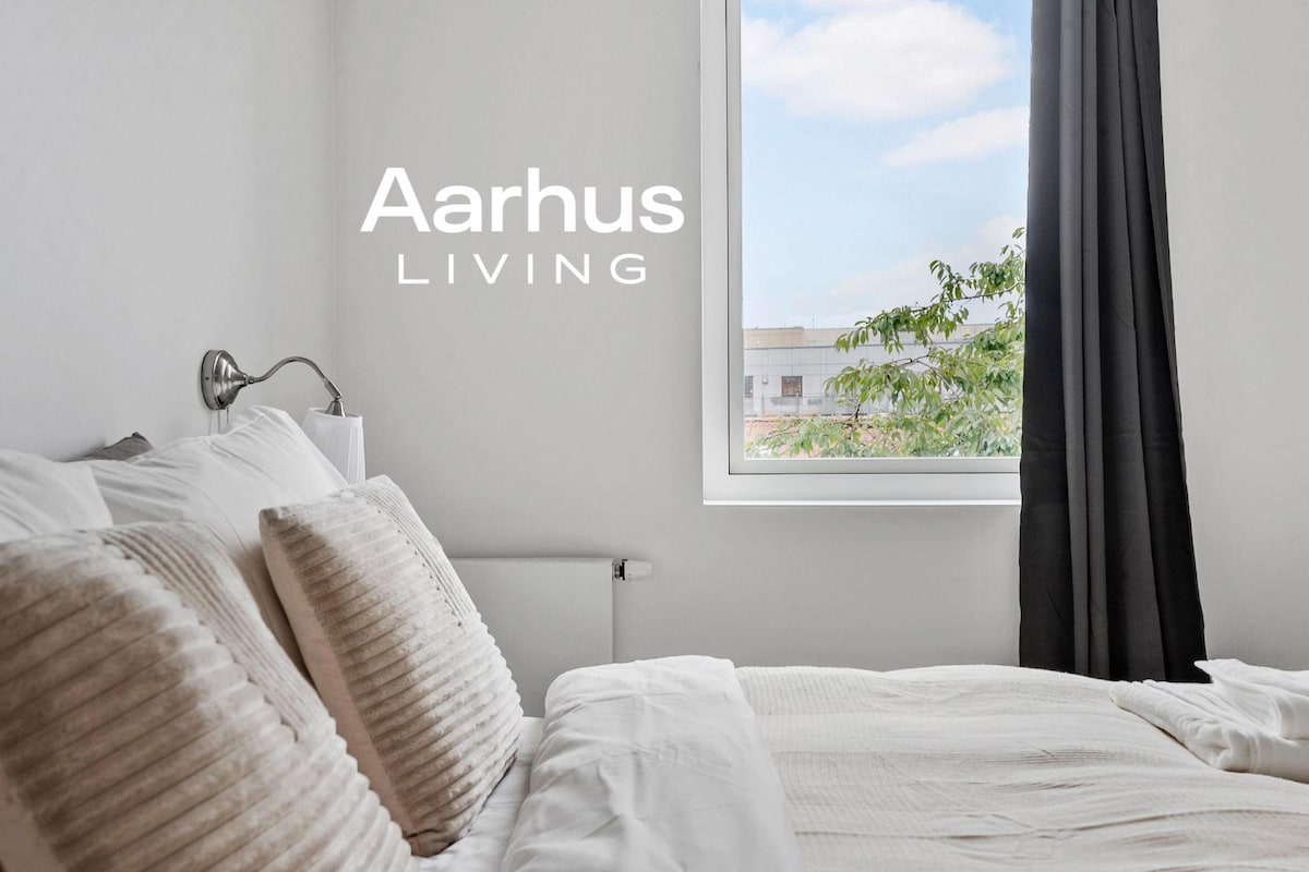 A bright bedroom features a spacious bed with plush pillows neatly arranged. Natural light filters through a large window, which is partially dressed with dark curtains. A sleek bedside lamp is mounted on the wall beside the bed, enhancing the room's minimalist design.