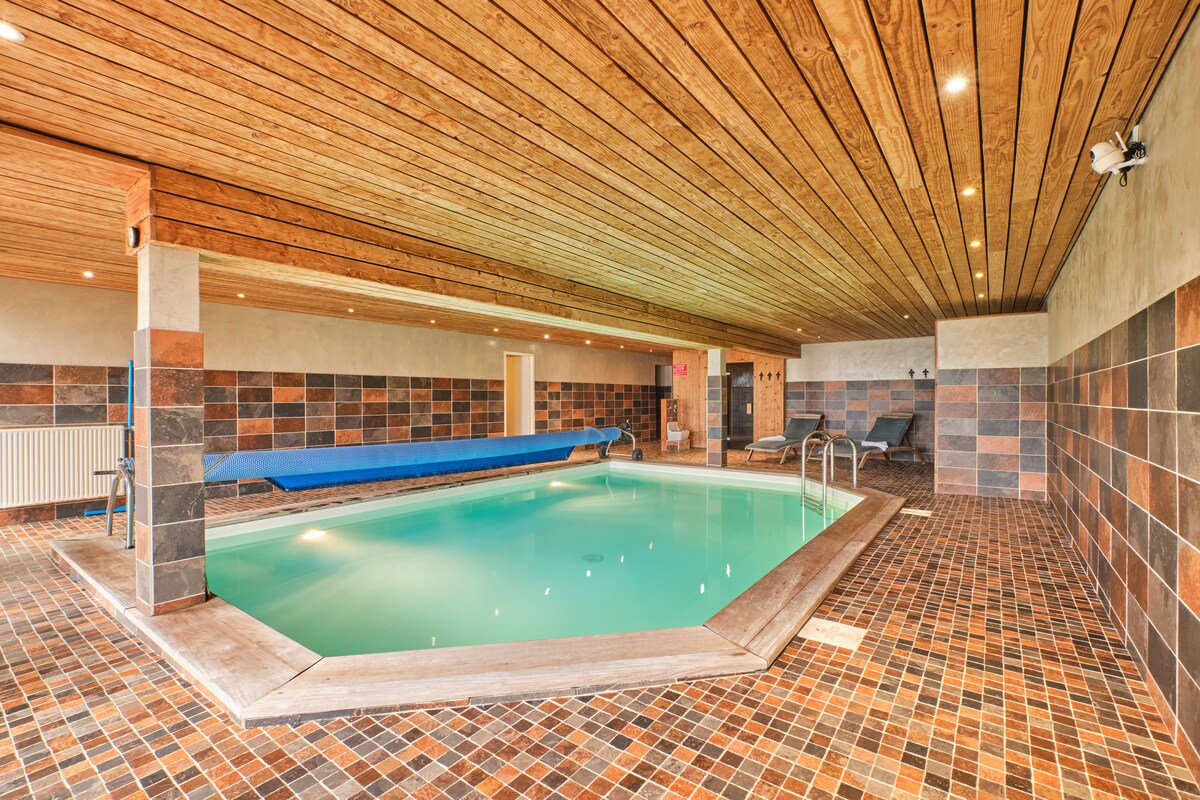An indoor swimming pool is surrounded by a tiled floor, featuring a wooden ceiling that enhances the space's warmth. Lounge chairs are positioned around the pool, and a long lap lane runs along one side. Soft lighting creates a serene atmosphere.