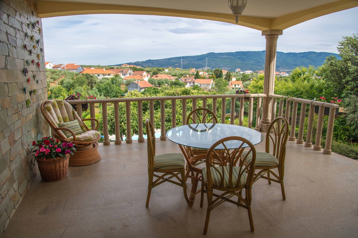 An open terrace is visible, featuring a glass table surrounded by four rattan chairs with green cushions. A comfortable armchair is positioned nearby. The view extends across a lush landscape, showcasing a mix of residential rooftops and distant mountains under a partly cloudy sky.