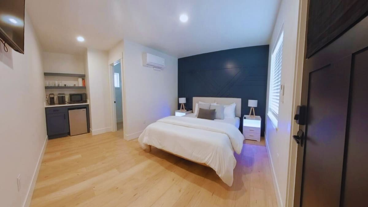 A well-lit bedroom features a cozy full-sized bed with crisp white linens and decorative pillows. Two bedside tables flank the bed, each with a lamp. In the background, a kitchenette is visible, equipped with a mini fridge, coffee maker, and microwave, enhancing functionality.