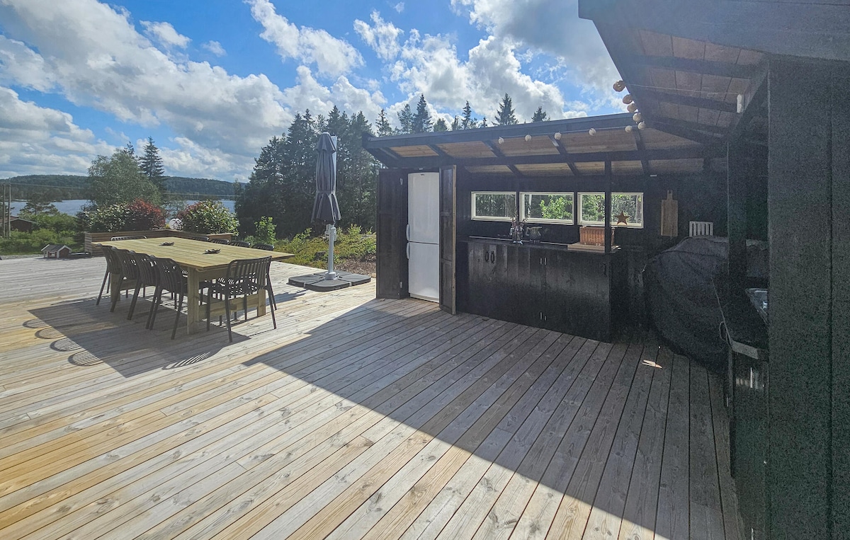An outdoor kitchen area features a spacious wooden deck with a dining table and chairs. A grill is visible, along with a sleek countertop and sink. The surroundings include greenery and trees, with views of the lake under a partly cloudy sky.