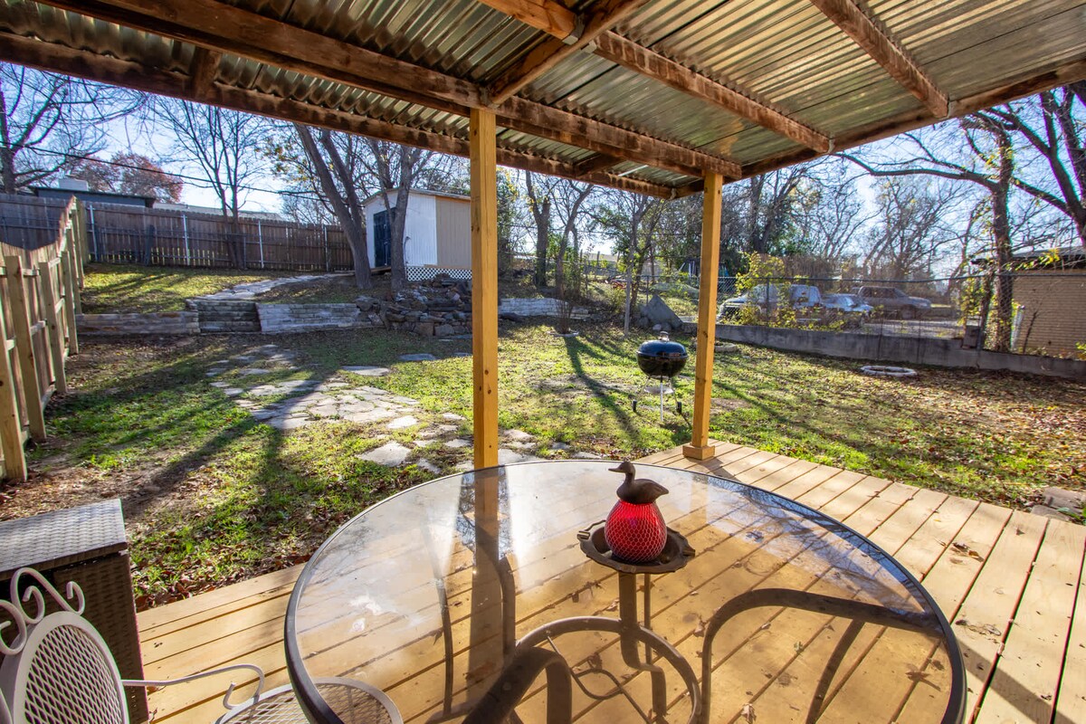 An outdoor covered deck features a circular table with transparent glass top and four surrounding chairs. A charcoal barbecue grill is positioned nearby. The spacious backyard is framed by trees, with a stone path leading to a distant shed, creating a serene outdoor setting.
