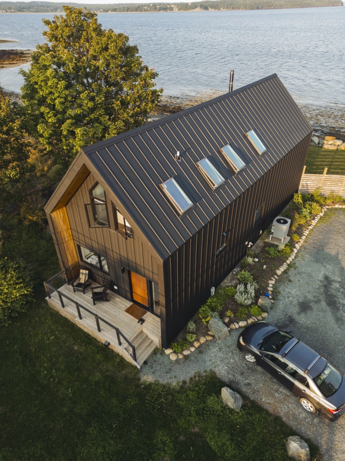 An aerial view showcases a modern Scandinavian-style retreat along the coastline. The dark exterior contrasts with the lush greenery surrounding the property. A spacious deck is visible in the front, complemented by a pathway leading to the entrance. Nearby, a vehicle is parked on a gravel area.