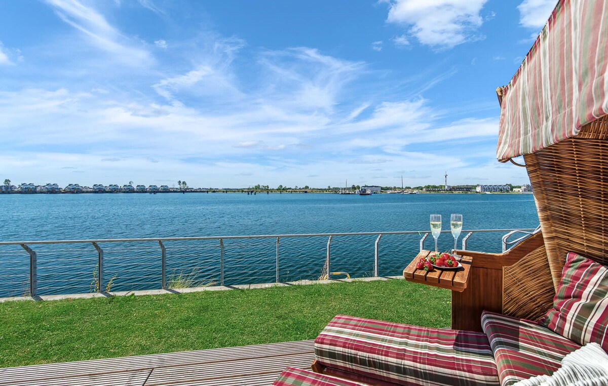 A comfortable beach chair is positioned on a grassy area overlooking the water, with glasses of sparkling beverage and strawberries placed on the side table. The calm water reflects the blue sky and fluffy clouds, creating a tranquil setting for relaxation.