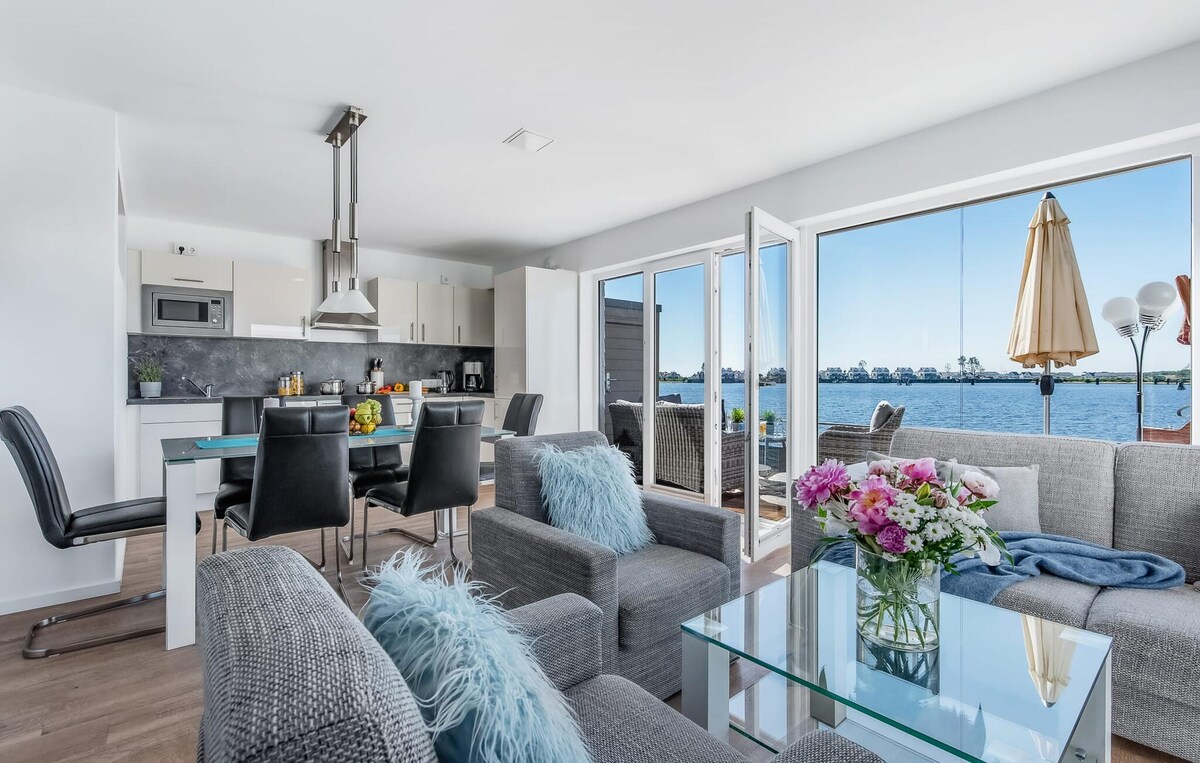 An open-plan living and dining area showcases contemporary furnishings, including a comfortable sofa and glass coffee tables. Floor-to-ceiling windows offer a view of the water. A dining table is set for meals, and a modern kitchen is visible in the background.