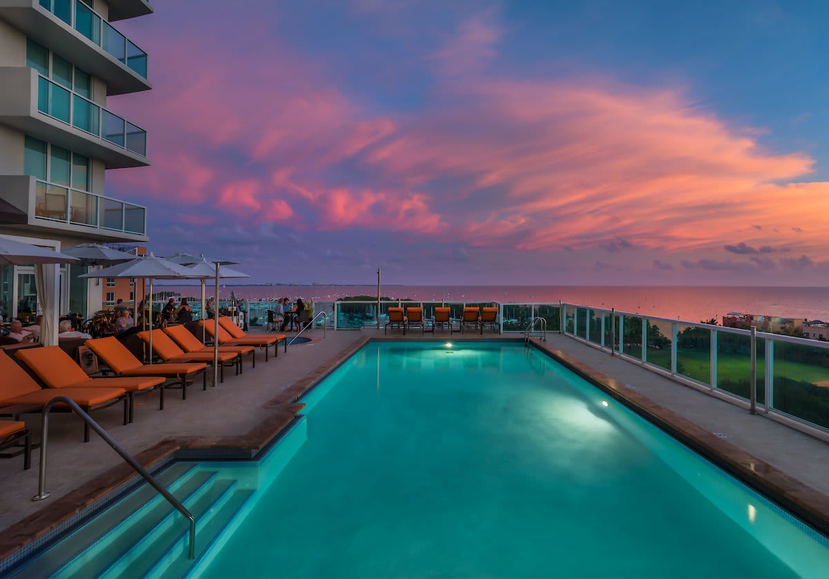An outdoor rooftop pool is framed by lounge chairs, providing a spacious area for relaxation. A vibrant sunset paints the sky in shades of pink and purple, enhancing the inviting atmosphere. The pool's pristine water reflects the colorful sky, creating a serene environment.