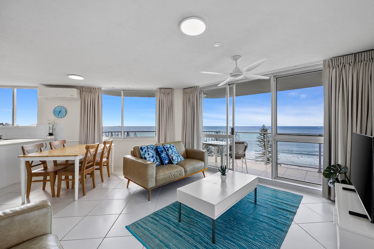 An open living area is presented with two large windows, allowing natural light and ocean views. A comfortable sofa faces a white coffee table, while a dining table with wooden chairs is positioned adjacent. A ceiling fan and light fixtures are included, enhancing the space.