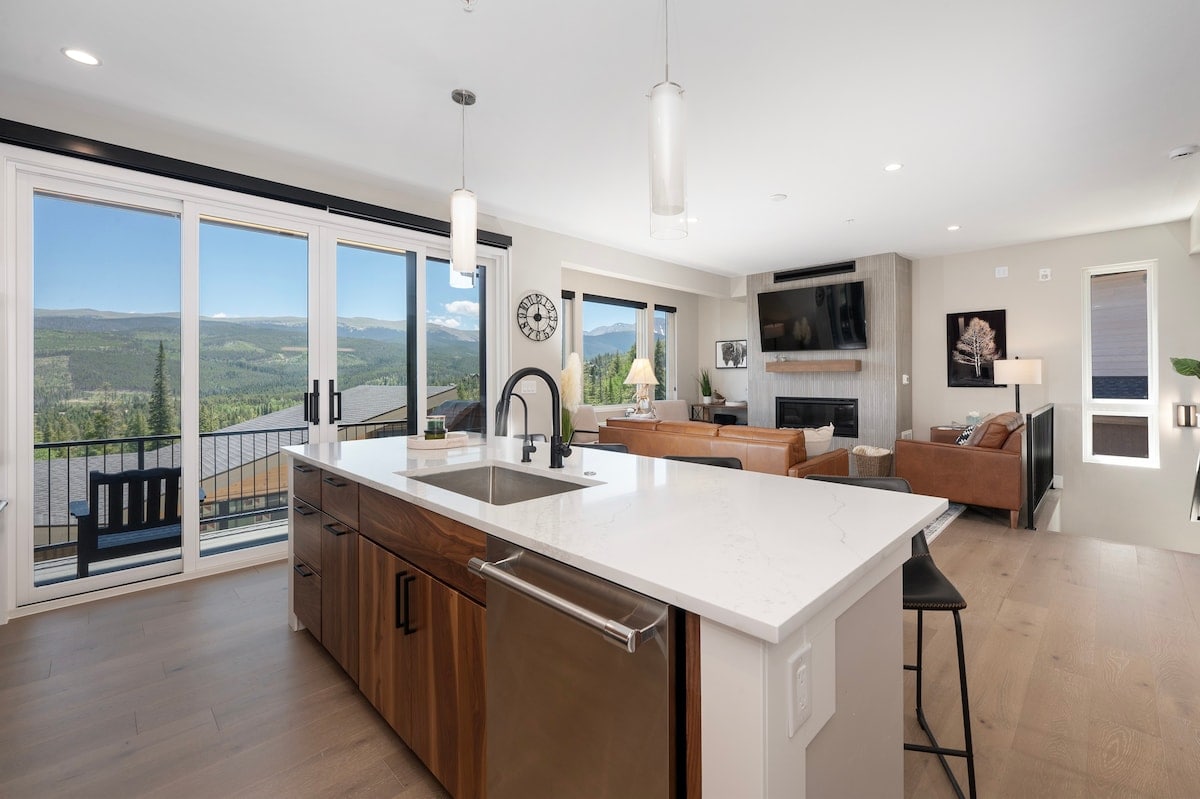 An open kitchen area features a large island that includes modern fixtures and a sleek design. Large glass doors allow for natural light and showcase stunning mountain views. The adjacent living space is furnished with comfortable seating and a modern fireplace.