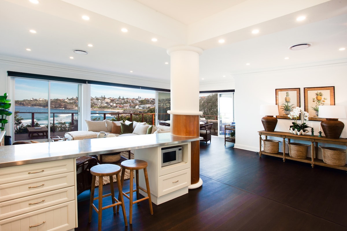 An open-plan living area is revealed, featuring a sleek kitchen island with bar stools. Sunlight fills the space, highlighting the elegant furnishings. Large windows frame views of the ocean and coastline, while decorative elements and a warm color palette create a welcoming environment.