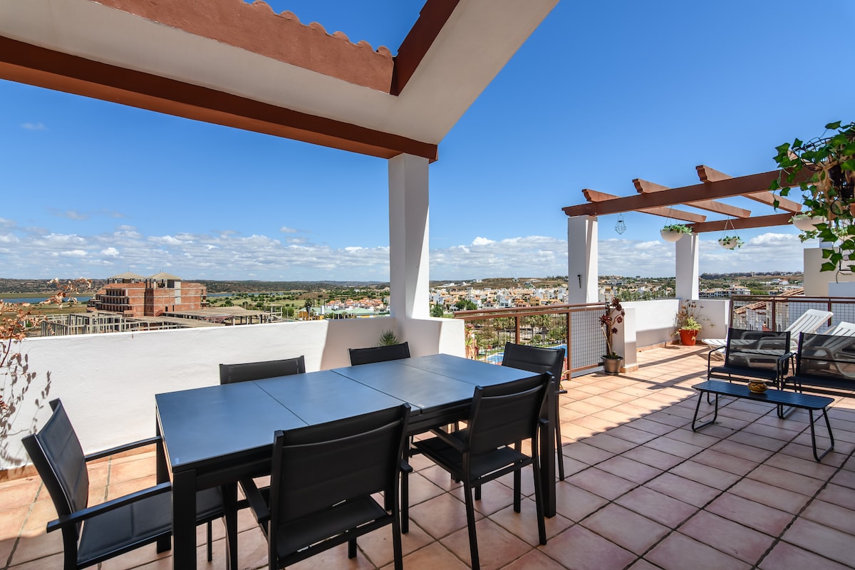 An outdoor terrace offers a dining area with a large table and black chairs, surrounded by scenic views. A shaded pergola adds comfort, while potted plants enhance the space. The tiled flooring reflects natural light, creating a serene environment for relaxation.