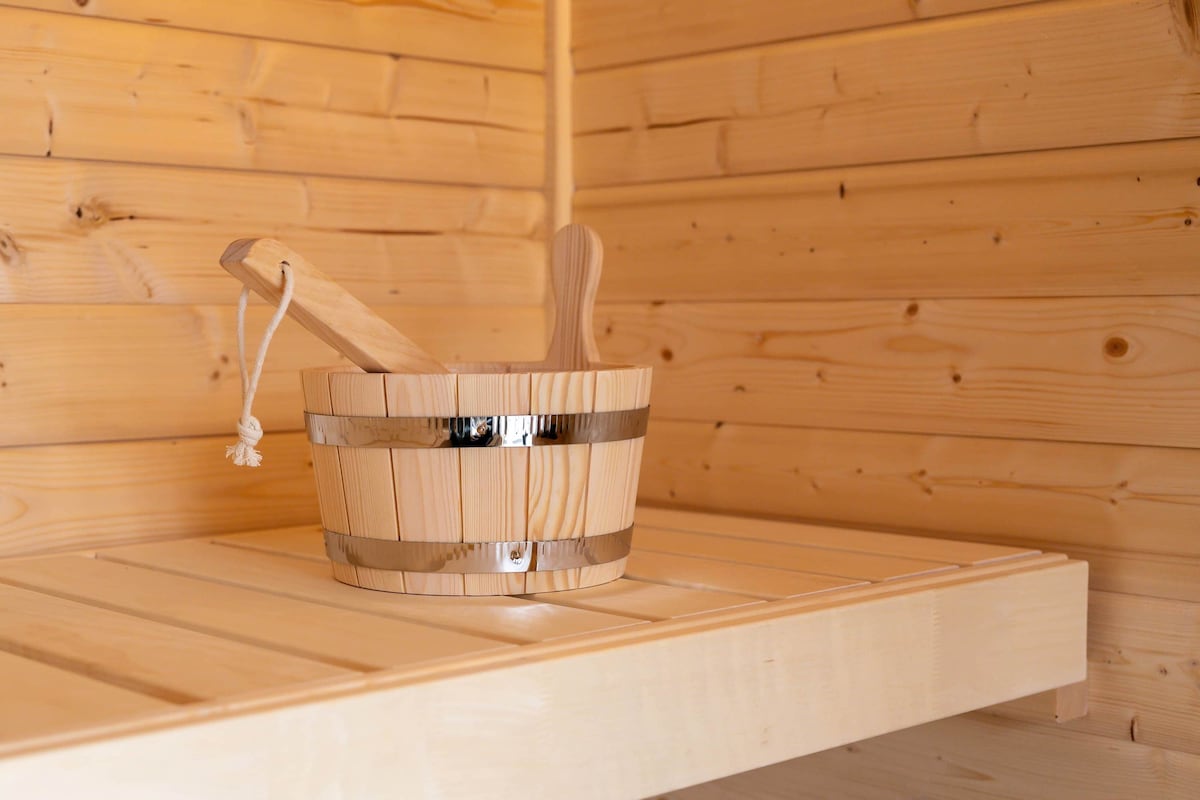 Inside the sauna, wooden paneling creates a warm environment. A wooden bucket with a metal band and two paddles are positioned on a bench made of smooth wood, highlighting a simple and functional design.