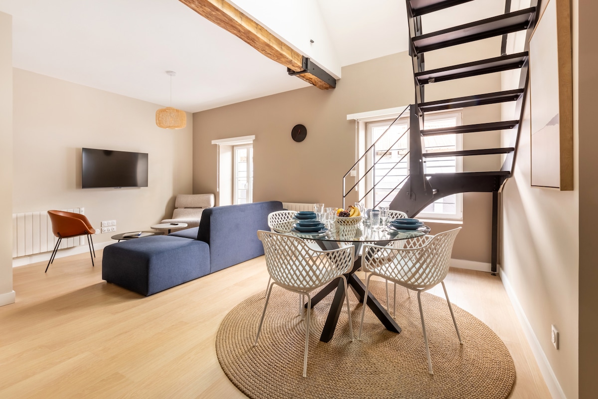 A modern living space features a spiral staircase leading to a loft area. A round dining table is set for four, with light-colored chairs surrounding it. Neutral tones are showcased in the walls and furniture, complemented by natural light flowing through the windows.
