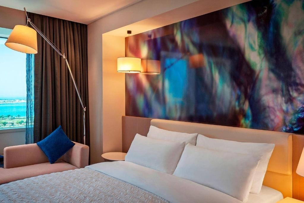 A chic guestroom features a spacious bed with white linens and a soft throw. An accent chair and a stylish floor lamp add comfort, while an abstract wall art creates a colorful focal point. A window offers views of the surrounding area.