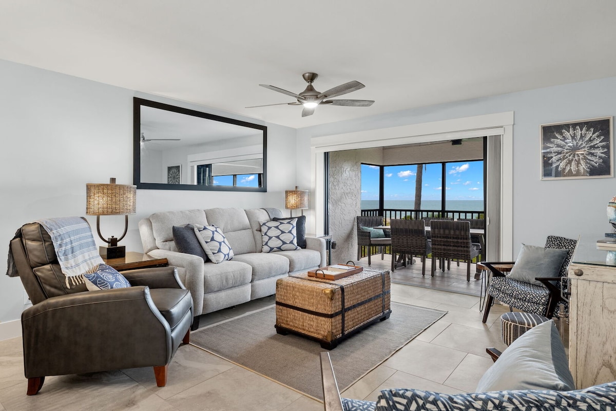 A bright living area is furnished with a comfortable gray sofa and accent chairs. A large mirror reflects natural light, while sliding glass doors provide access to a balcony with water views. Decorative elements include a coffee table and stylish lamps.