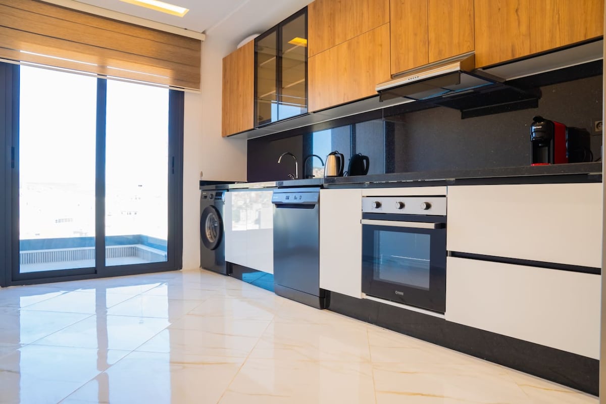 The modern kitchen features sleek cabinetry in warm wood tones, complemented by glossy black countertops. Stainless steel appliances, including a refrigerator and oven, are visible. Large windows allow natural light to fill the space, while a washing machine and coffee maker are conveniently placed.