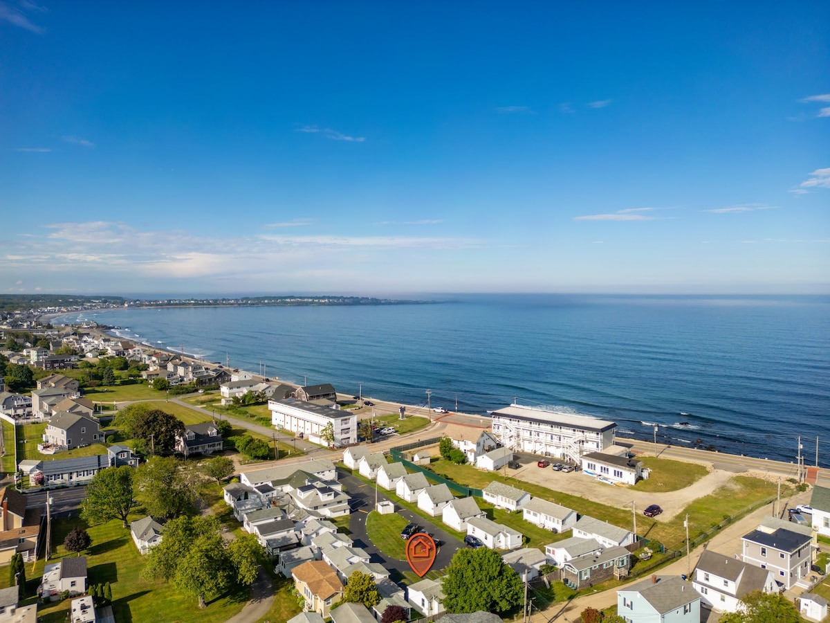 An aerial view captures the coastline of York, showcasing long stretches of beach and gently rolling waves. Nearby structures are visible, including residential homes and commercial buildings, set against a backdrop of clear blue skies that complement the tranquil sea.