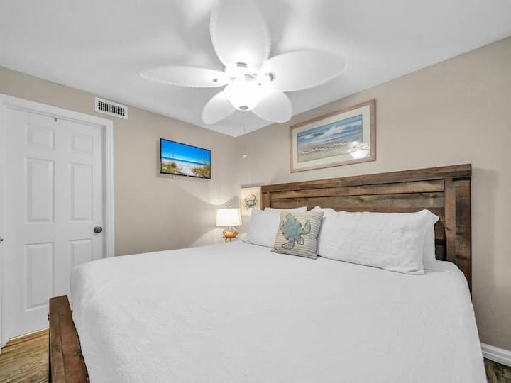 Pumzika kwa utulivu katika chumba chako cha kulala chenye mandhari ya pwani kilicho na kitanda chenye kichwa cha mbao za asili, mashuka safi na upepo mwanana wa feni ya dari.