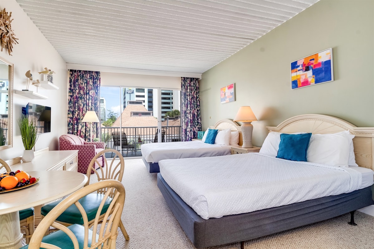 A spacious studio suite features two queen beds with white linens and decorative pillows. A cozy seating area includes a patterned chair and a circular dining table. Bright artwork adorns the walls, while large windows with floral curtains allow natural light to fill the room.