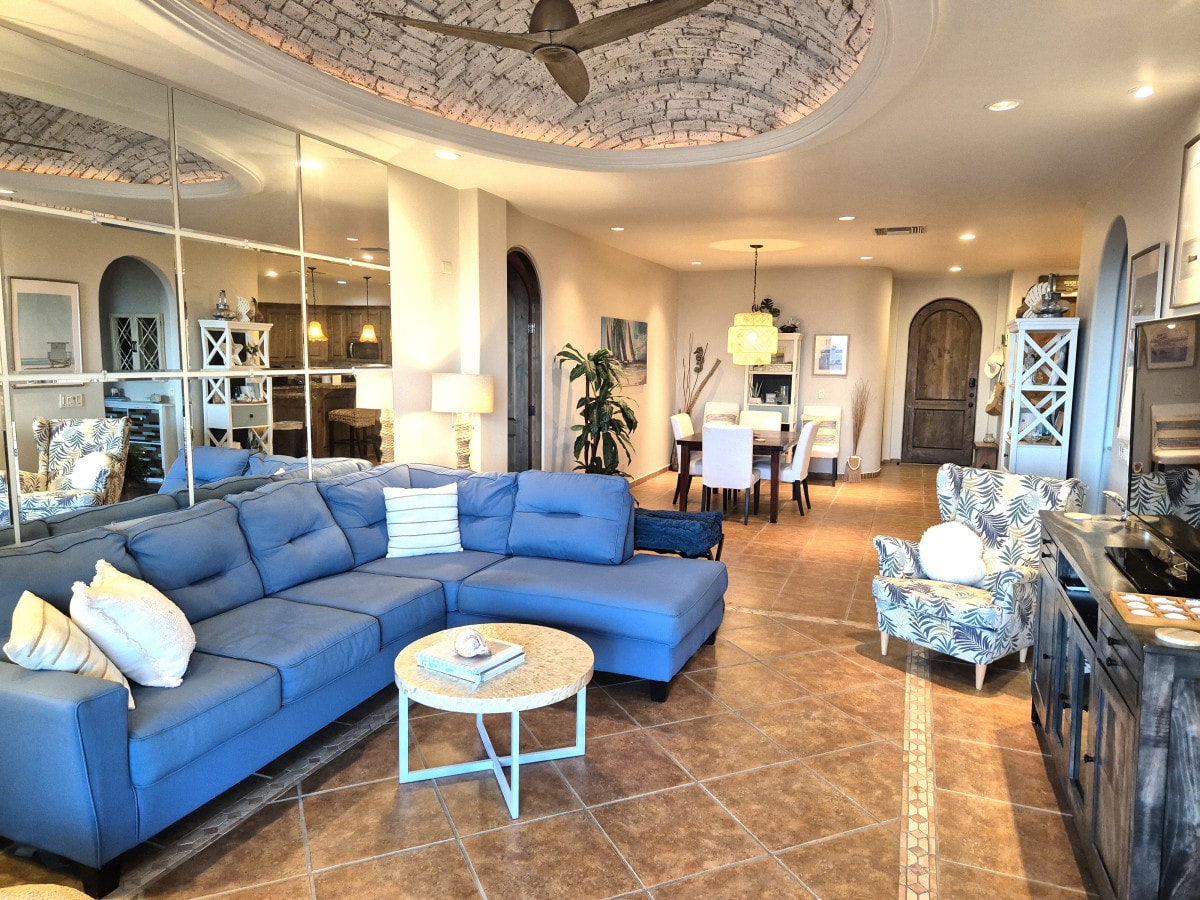 A spacious living area features a blue sectional sofa and an accent chair with a floral pattern. A circular coffee table sits in front, and large mirrors reflect the open space. The dining area is visible in the background, highlighting a neutral palette and light fixtures.