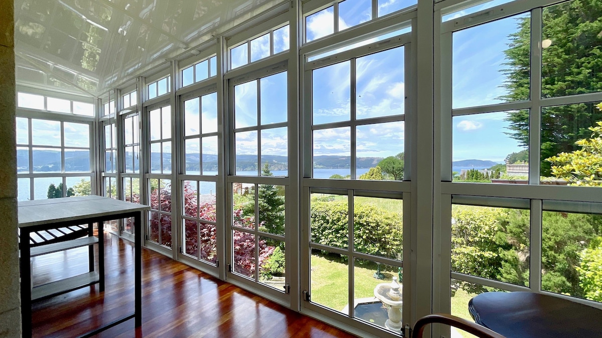 A sunlit indoor space is framed by large glass windows, offering panoramic views of the tranquil waters and lush gardens outside. The gleaming wooden floor is visible, along with a simple table positioned to maximize the scenic outlook.