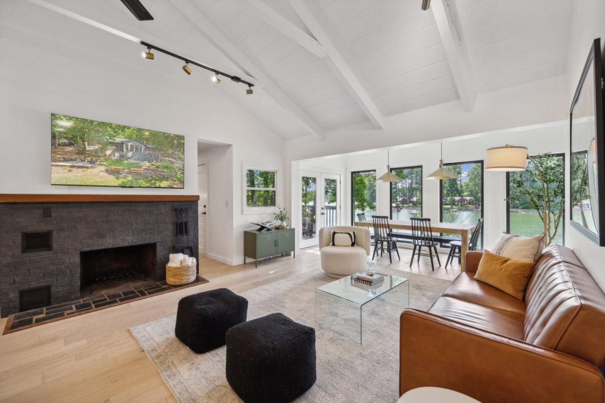 An open concept living room features soaring ceilings and large windows that provide views of the outdoor greenery. A cozy fireplace is set against a black accent wall, complemented by a sleek glass coffee table and comfortable seating. A dining area is visible through the windows.