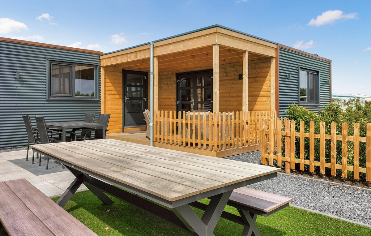 A modern holiday home exterior is showcased, featuring a covered wooden terrace at the entrance. A spacious dining table is positioned in the foreground, with comfortable seating visible. Surrounding greenery and a wooden fence offer a welcoming outdoor environment.