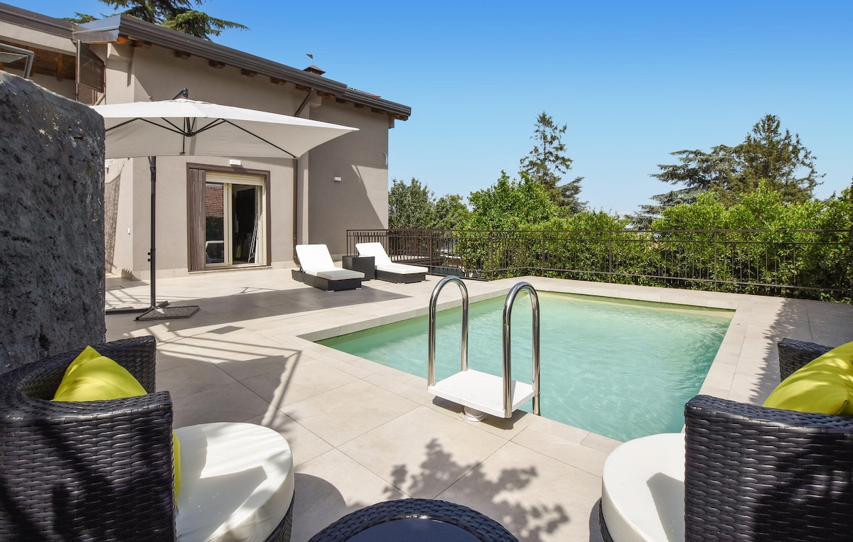 A serene outdoor area features a private pool surrounded by a tiled deck. Two sun loungers are positioned beside the pool under a large umbrella. A circular seating arrangement with light cushions is evident, along with well-maintained greenery providing a sense of privacy.
