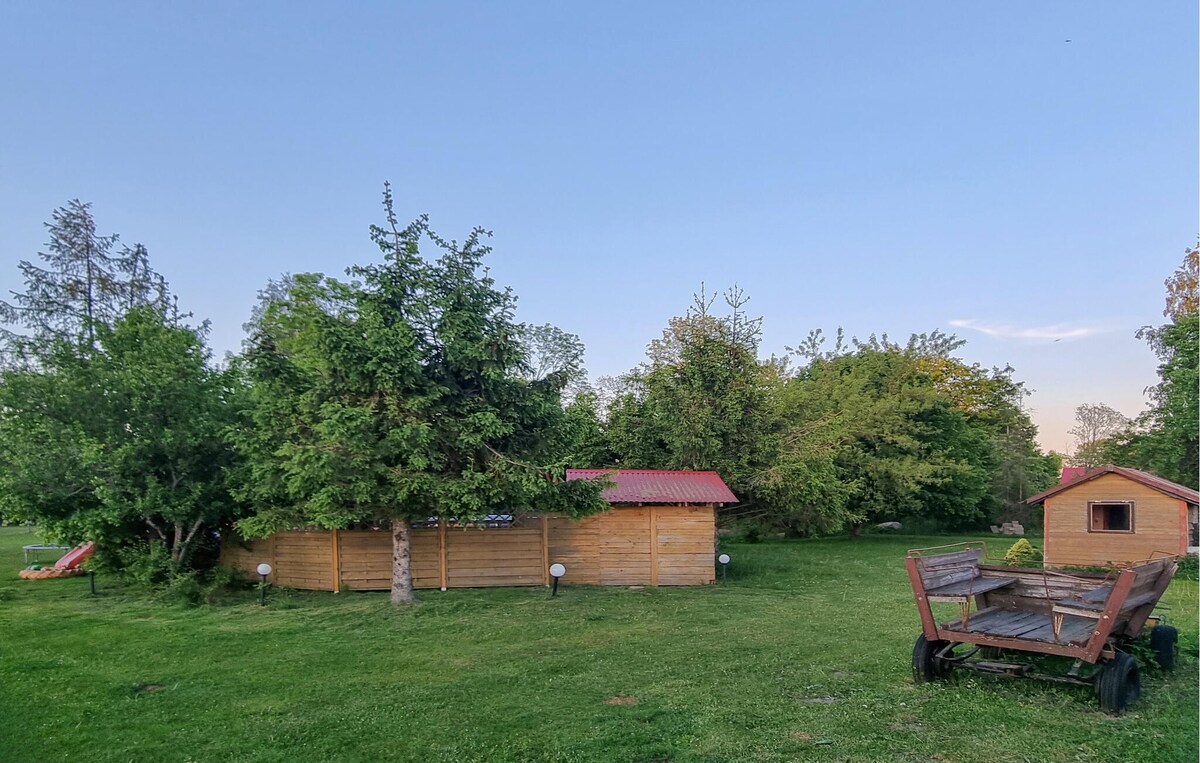 A spacious outdoor area is surrounded by lush greenery, featuring wooden structures with red roofs. A rustic wooden cart is placed on the grass, alongside well-maintained trees and shrubs. Soft lighting from ground fixtures is visible, enhancing the natural setting.