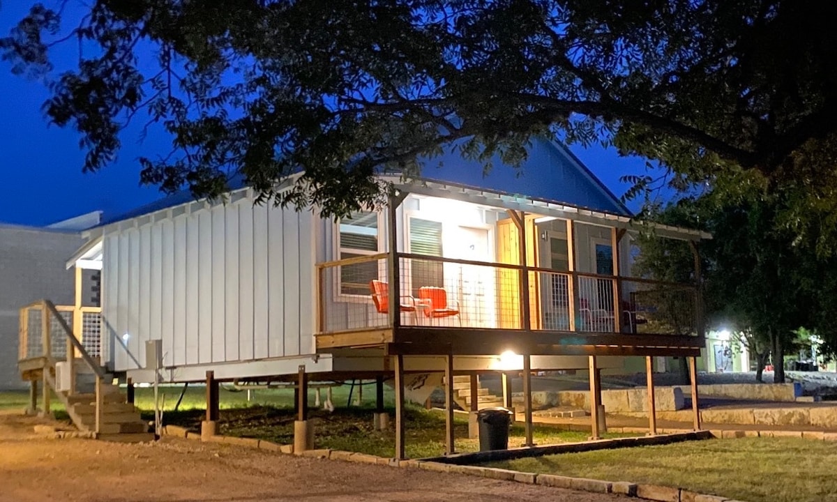 A newly constructed cabin is viewed at dusk, showcasing a welcoming porch with two bright red chairs. The structure is elevated with steps leading up to the entrance. Soft lighting illuminates the front, highlighting the minimalist design and surrounding grassy area.