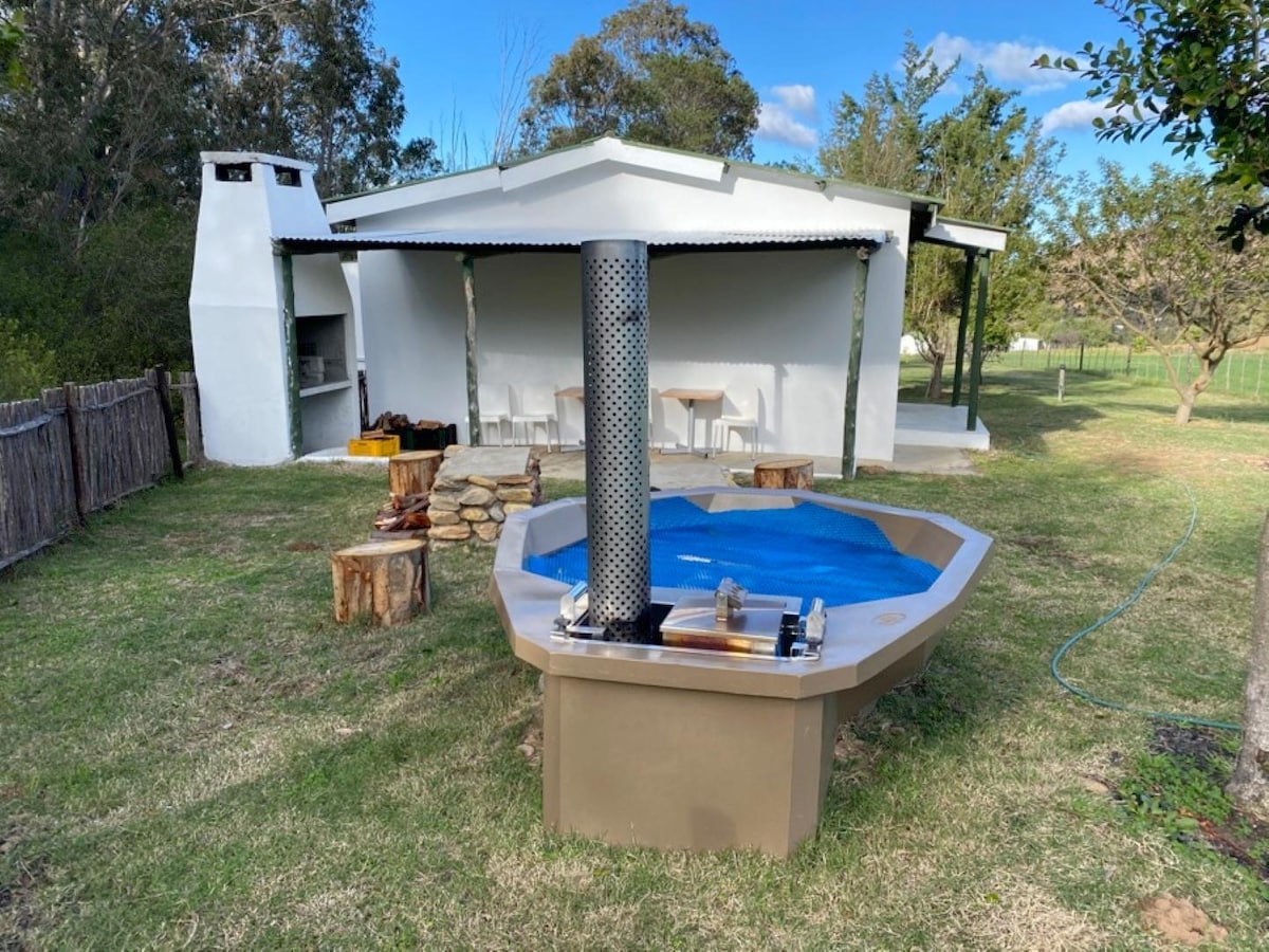 An outdoor area features a unique hot tub situated on a grassy lawn, surrounded by trees. A barbecue area with a stone grill stands nearby, shaded by a roof. A simple table with chairs is visible in the background, complemented by a peaceful natural setting.