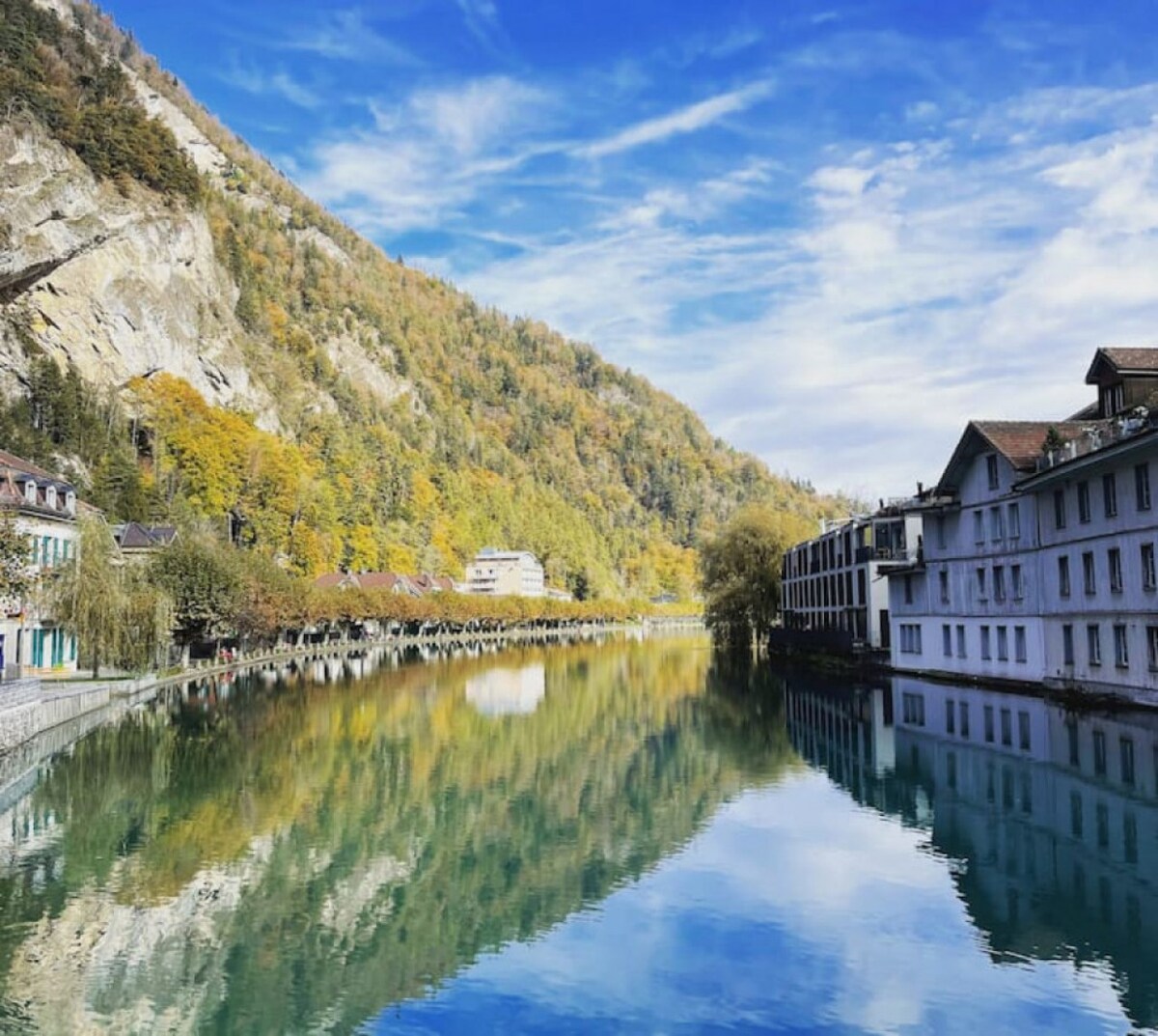 A serene river reflects the surrounding autumn foliage and nearby buildings. The mountains rise in the background, creating a picturesque landscape under a bright blue sky with light clouds. The calm water captures the vibrant colors of the trees, adding to the tranquil ambiance.