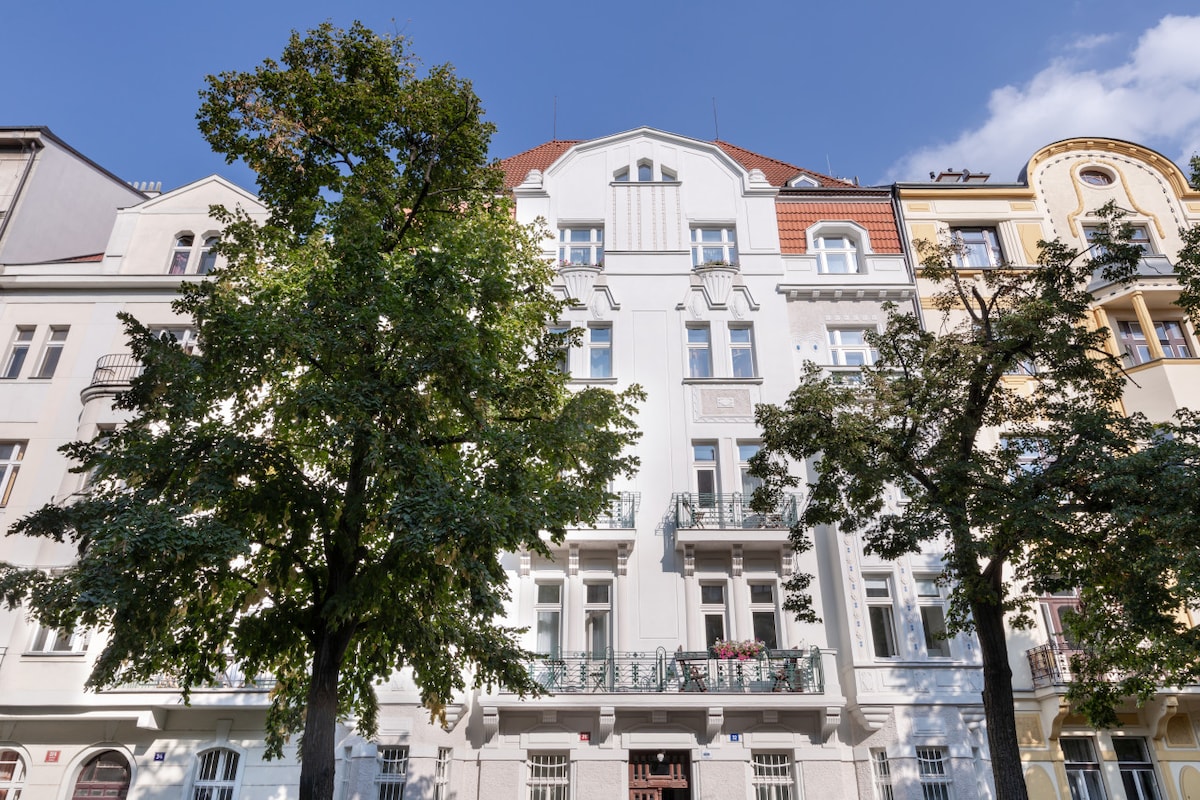 The exterior of a classic building showcases a white façade detailed with decorative elements. Symmetrical windows with balconies are visible on both upper and lower levels. Lush green trees frame the structure, enhancing the charm of the surrounding neighborhood.