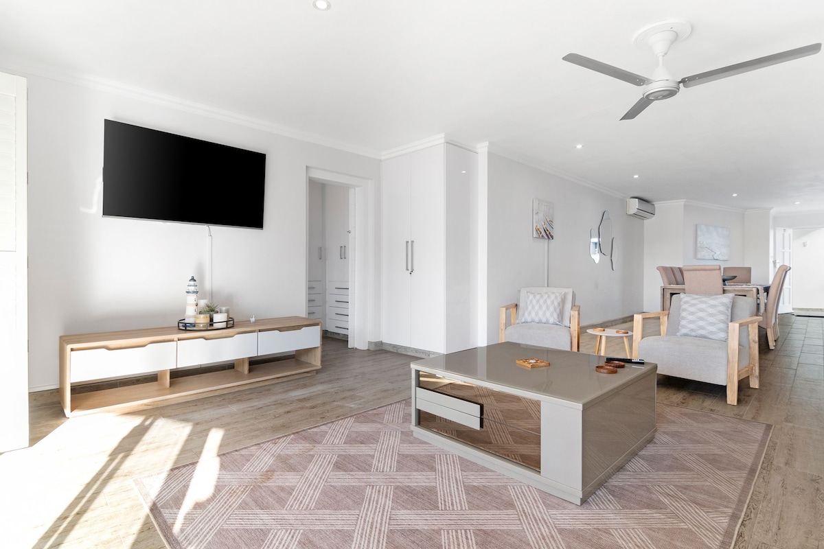 A spacious lounge area is showcased with light-colored furnishings and an elegantly designed coffee table. A large television is mounted on the wall opposite. Natural light floods the room, illuminating the wood-like flooring that extends to a dining area in the background.