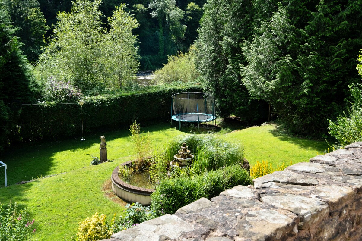 A spacious garden features a well-maintained lawn, surrounded by dense greenery. A trampoline is positioned in one corner, while a circular pond with a stone fountain sits near the center. Sunlight filters through the trees, creating a tranquil outdoor atmosphere.