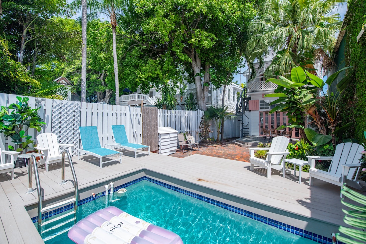 A private outdoor space is highlighted by a swimming pool surrounded by lounge chairs and tropical greenery. A wooden deck features a shaded area with additional seating, creating a tranquil retreat. The pool area is enclosed by a white fence, ensuring a sense of privacy.