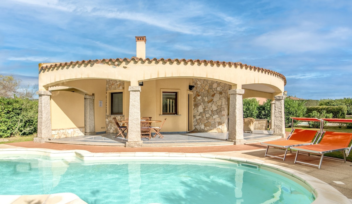 An outdoor space features a round covered patio with stone columns, overlooking a private pool. Lounge chairs are positioned nearby, and lush greenery provides privacy. Large windows allow for natural light to enter the interior space, enhancing the serene environment.