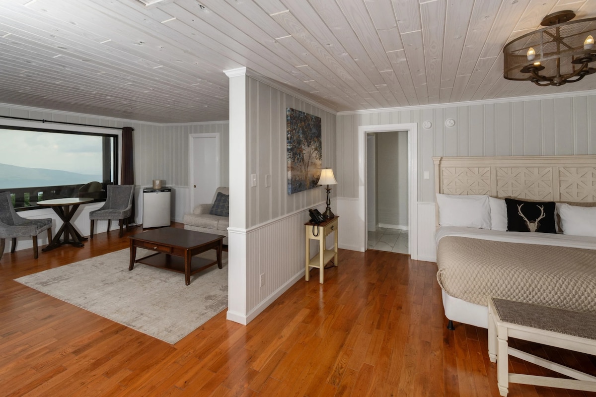 An open suite presents a seating area with two chairs by a round table and a small side table. A spacious bed is placed against a textured wall, with natural light filtering through large windows, showcasing views of the surrounding mountains.