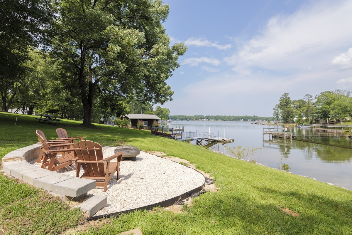 A cozy seating area with wooden chairs is situated on a pebble stone surface, facing the calm waters of the lake. Lush green grass surrounds the area, providing a natural and serene setting. The private dock is visible in the distance, offering access to the water.