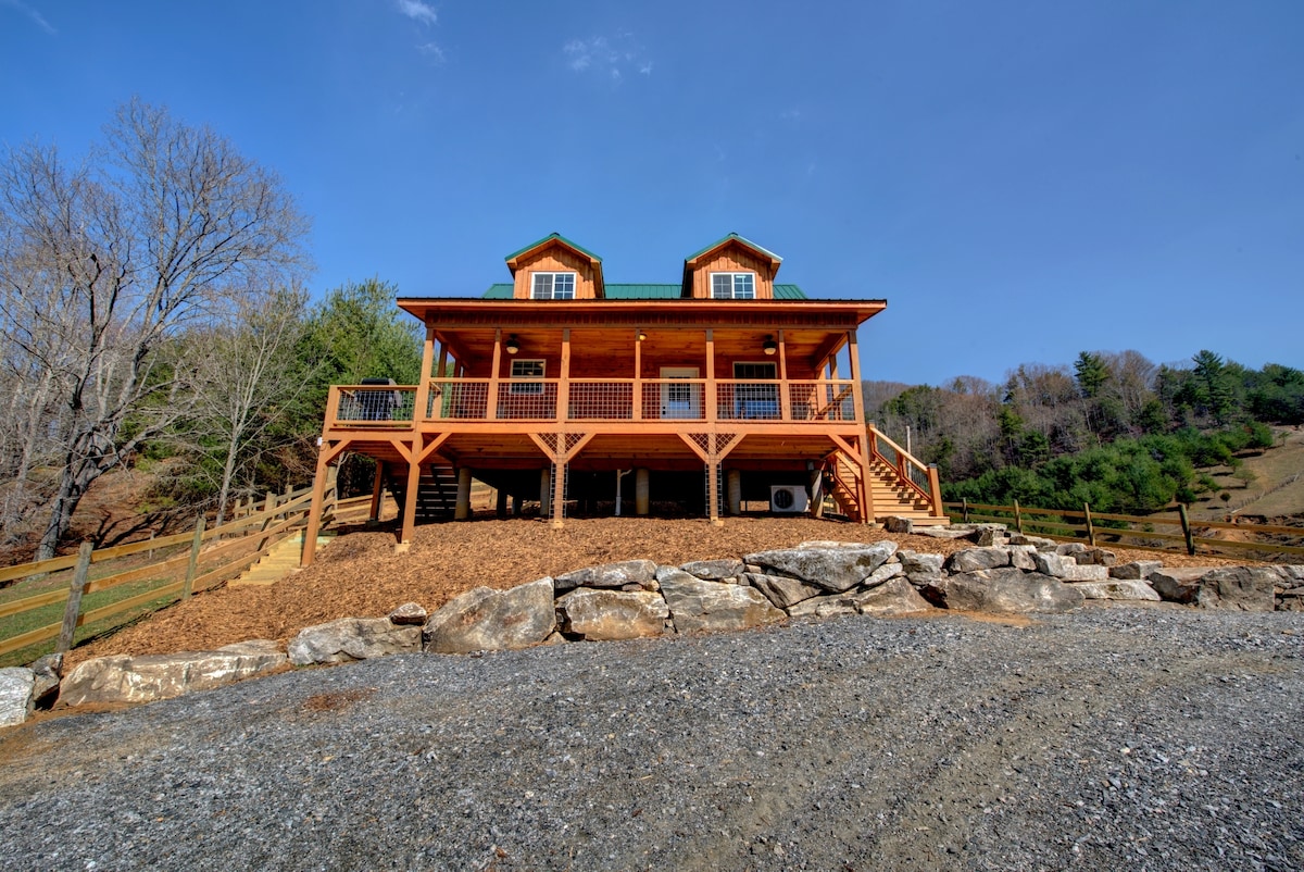 The two-story log cabin is set on a gentle slope, featuring a wraparound porch supported by wooden posts. Large windows illuminate the interior, while a green metal roof contrasts with the natural wooden exterior. Surrounding landscapes of trees and rolling hills are visible in the background.